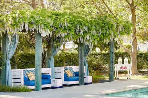 Poolside lounge area with blue and white daybed under teal pergola draped in sheer curtains beneath blooming wisteria trees