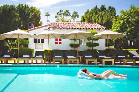 Woman floating in turquoise pool with Spanish-style white building and umbrellas in background