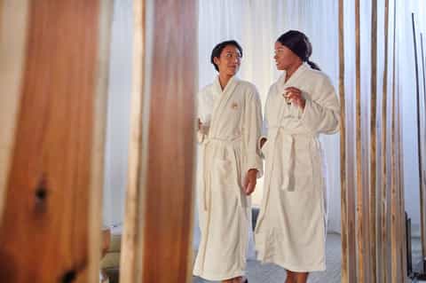 Two women in cream-colored robes conversing in a bright modern spa or hotel hallway