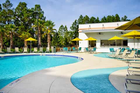 Resort pool with curved design, yellow umbrellas, palm trees, and white clubhouse building