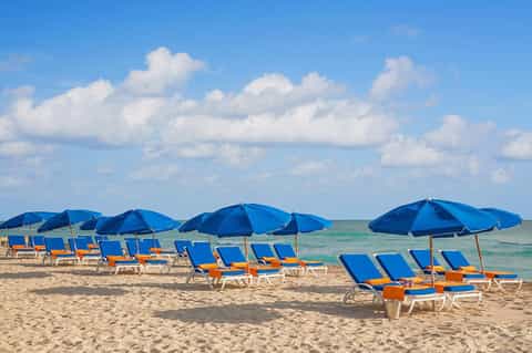 Sandy beach with rows of blue umbrellas, orange lounge chairs, turquoise ocean, and cloudy sky