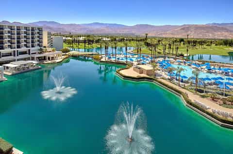 Resort aerial view with multiple pools, palm trees, mountains, and blue lounge chairs
