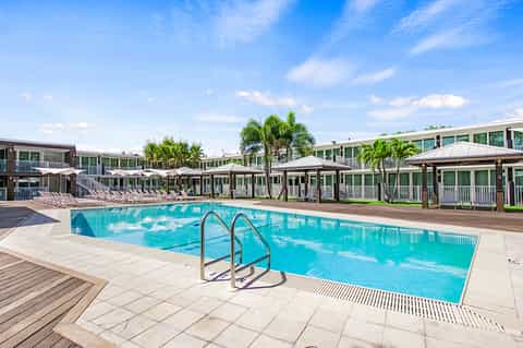 Resort pool with family lounge area, white umbrellas, palm trees, and green cottage-style buildings