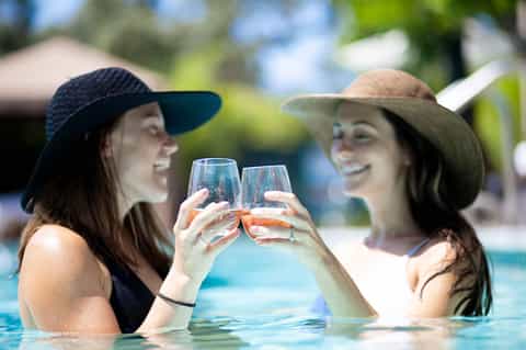 Two women toasting with drinks while in resort pool, wearing sun hats on sunny day