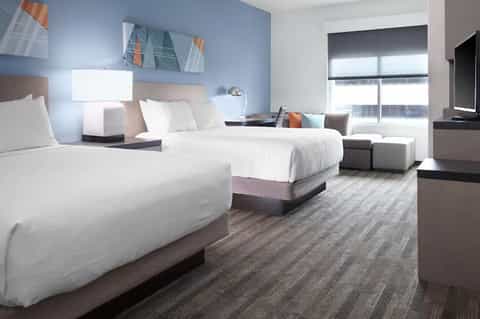 Modern hotel guestroom with two queen beds, blue accent wall, TV, and floor-to-ceiling windows