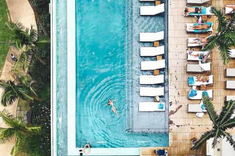 Aerial view of resort pool with white cabanas and wooden deck surrounded by palm trees and beach
