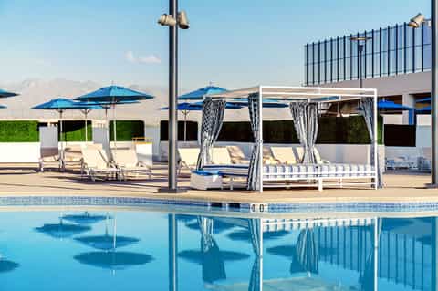 Resort poolside with blue umbrellas, white cabanas with curtains, lounge chairs, and mountain backdrop
