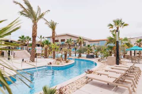 Resort pool area with curved lazy river, palm trees, lounge chairs, and main building in background