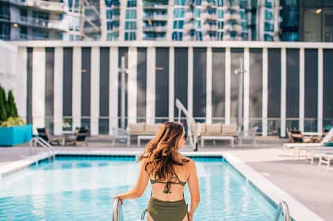 Woman climbing pool ladder in modern resort pool with high-rise buildings and white fencing