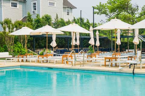 Resort pool with white umbrellas, wooden lounge chairs, pergolas, and manicured landscaping near residential building