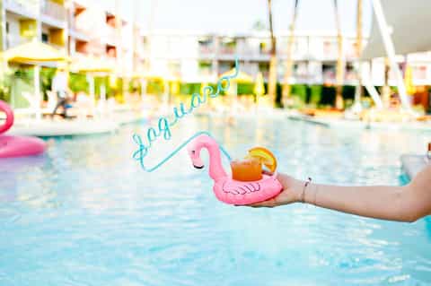 Hand holding pink flamingo pool float with cocktail by resort pool