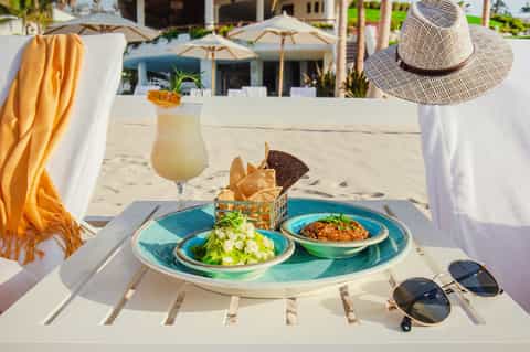 Beach dining setup with fresh salad, cocktail, chips, straw hat, and ocean view backdrop