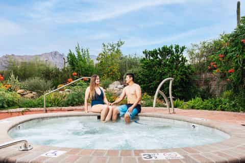 Couple sitting at edge of hot tub with desert mountains, flowering plants and vegetation visible in background