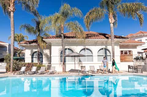 Resort pool with palm trees, white cabana building, and guests walking along pool deck