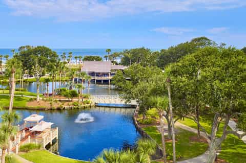 Aerial resort view with lagoon, fountain, thatched structures, palm trees, and ocean horizon