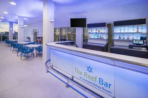 Modern hotel bar with blue lighting, liquor shelves, seating area, and TV screens