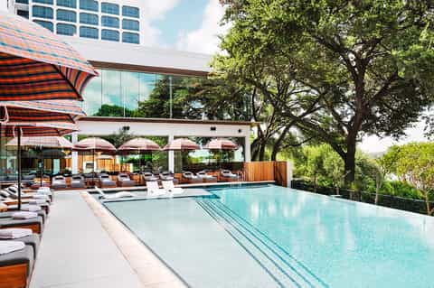 Luxury resort pool with blue water, lounge chairs, striped umbrellas, and tall trees under clear sky