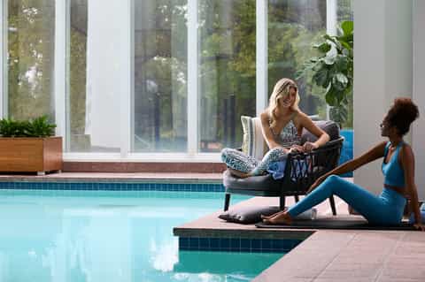 Two women relaxing by indoor pool with large windows overlooking forest scenery