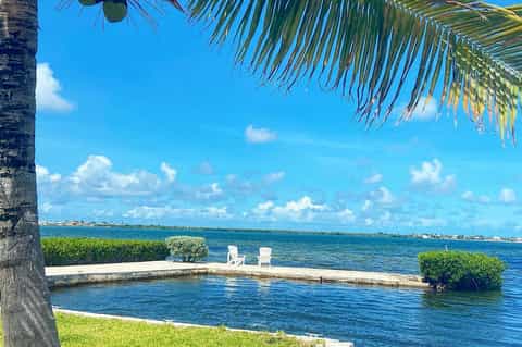 Waterfront seating area with white Adirondack chairs under palm fronds and clear blue sky