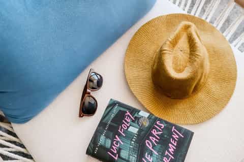 Poolside flat lay with straw hat, sunglasses, book, and blue lounge chair beside sparkling pool water