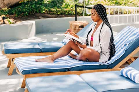 Woman reading on striped lounge chair in sunny resort pool area with hedges