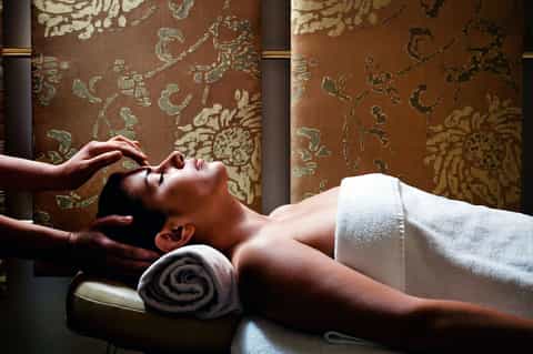 Spa treatment room with therapist giving facial massage, ornate gold wallpaper, and rolled towels