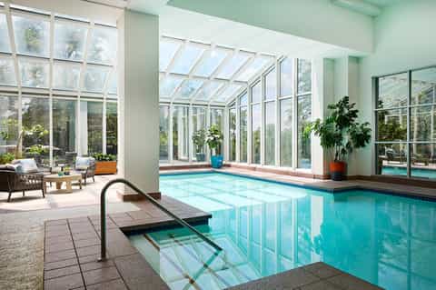 Indoor pool with glass roof, surrounding lounge seating, indoor plants, and natural light from windows