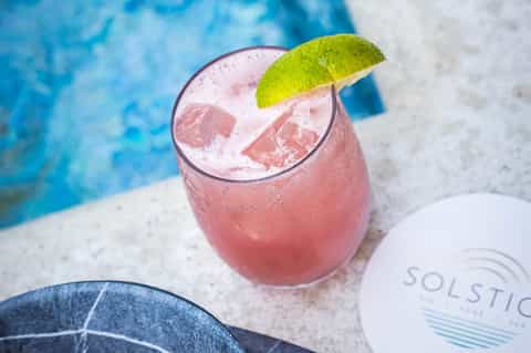 Pink cocktail with lime garnish on pool deck with turquoise water in background