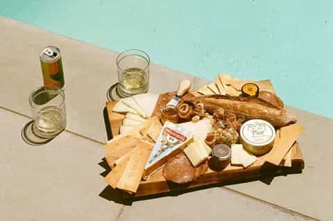 Poolside charcuterie board with cheeses, bread, and beverages beside turquoise water