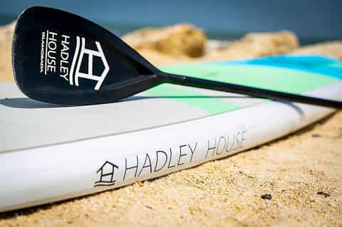 Paddle board with Hadley House logo on sandy beach near ocean rocks
