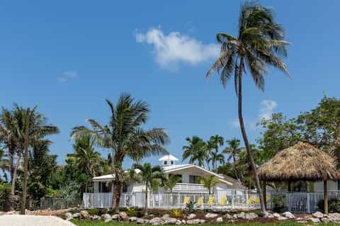 Beachfront tropical cottage with white railings, thatched-roof structure, and surrounding palm trees