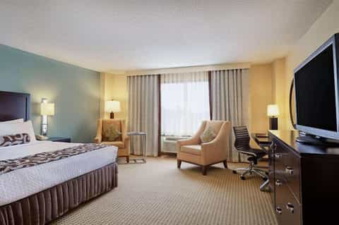 Hotel bedroom with queen bed, beige armchair, flat-screen TV, and window with drapes