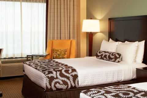 Modern hotel room with king bed, brown patterned bedding, orange chair, windows with curtains, and desk