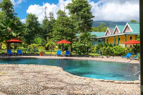 Tropical beach resort with colorful buildings, red umbrellas, blue lounge chairs, turquoise water, and lush green landscape