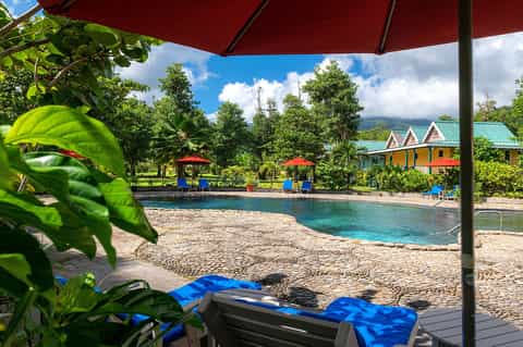 Tropical resort beach with colorful cabanas, turquoise water, lush vegetation, and red umbrella shade