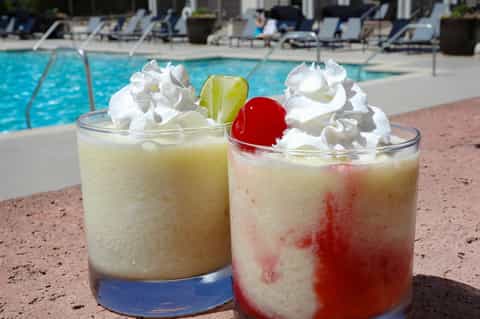 Two colorful tropical cocktails with whipped cream, cherries and lime garnishes poolside