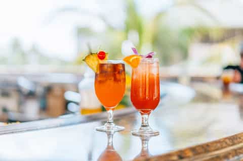Two tropical cocktails with fruit garnishes on a poolside bar counter