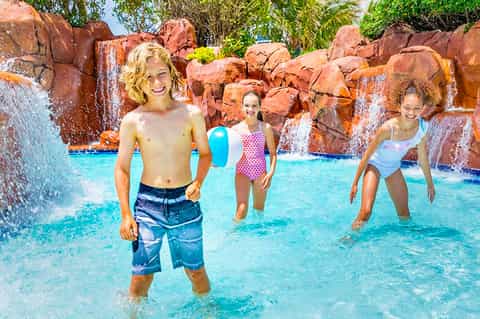 Children playing in a bright turquoise wave pool with red rock formations and waterfalls