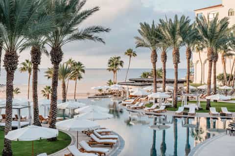 Luxury beachfront resort pool with palm trees, loungers, umbrellas, and ocean views