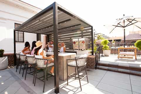Rooftop bar with pergola structure, patrons at counter, city skyline views, and umbrella seating