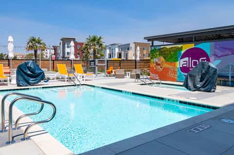 Modern resort pool with colorful mural, yellow loungers, and buildings visible beyond wooden fence