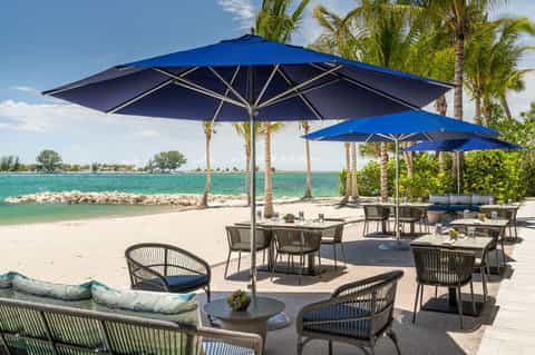 Beach club seating area with large blue umbrellas, wicker chairs, turquoise water, and palm trees