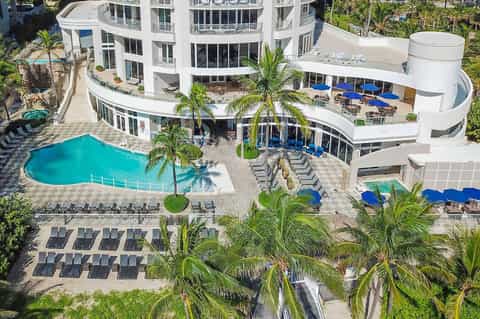 Aerial view of resort with curved white building, turquoise pool, palm trees, and blue umbrellas