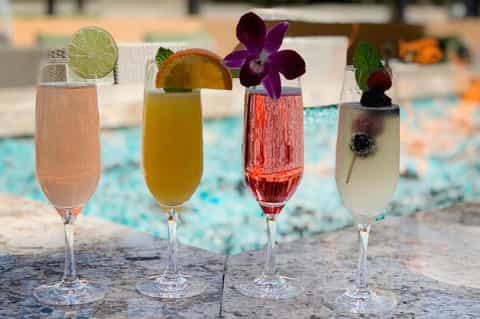 Four colorful cocktails with fruit garnishes and flowers by a resort pool