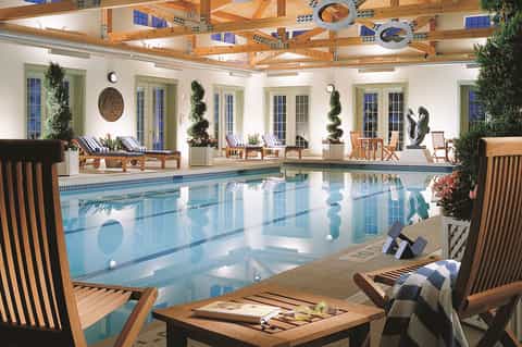 Indoor lap pool with wooden beam ceiling, floor-to-ceiling windows, lounge seating, and plant décor