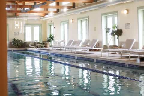 Indoor lap pool with lane dividers, lounge chairs, wood ceiling beams, and bright natural lighting