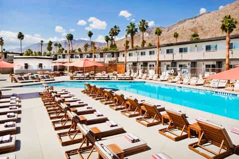 Resort pool with turquoise water, wooden lounge chairs, palm trees, and mountain backdrop