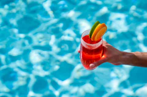 Hand holding red cocktail with sugared rim and fruit garnish over sparkling blue pool water