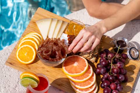 Poolside snack board with fresh fruit, cheese, and beverages on wooden serving platter