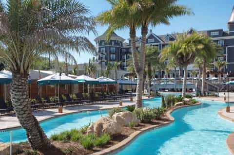 Lazy river pool at waterfront resort with tropical palm trees, umbrellas, multi-story hotel buildings, and boat dock